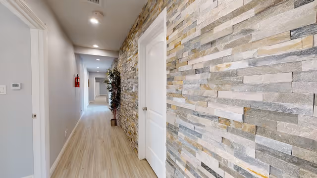 A well-lit interior hallway with light wood flooring and a textured stone accent wall on the right. There are white doors along the hallway and a potted plant placed near the middle. The ceiling has recessed lighting and a fire extinguisher is mounted on the left wall.