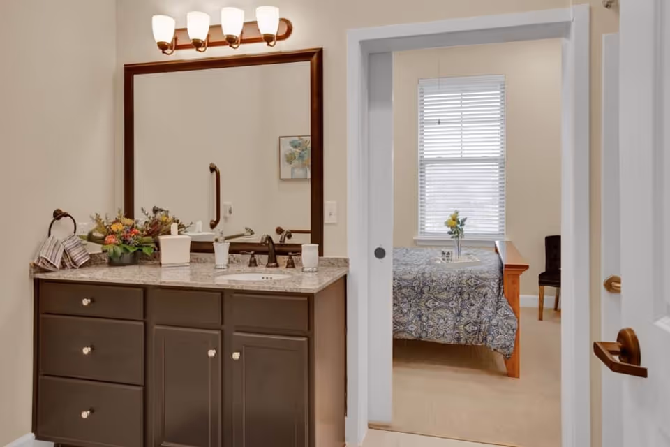 Bathroom vanity with a granite countertop, mirror and light fixture opening through a sliding door into a bedroom with a bed and window.