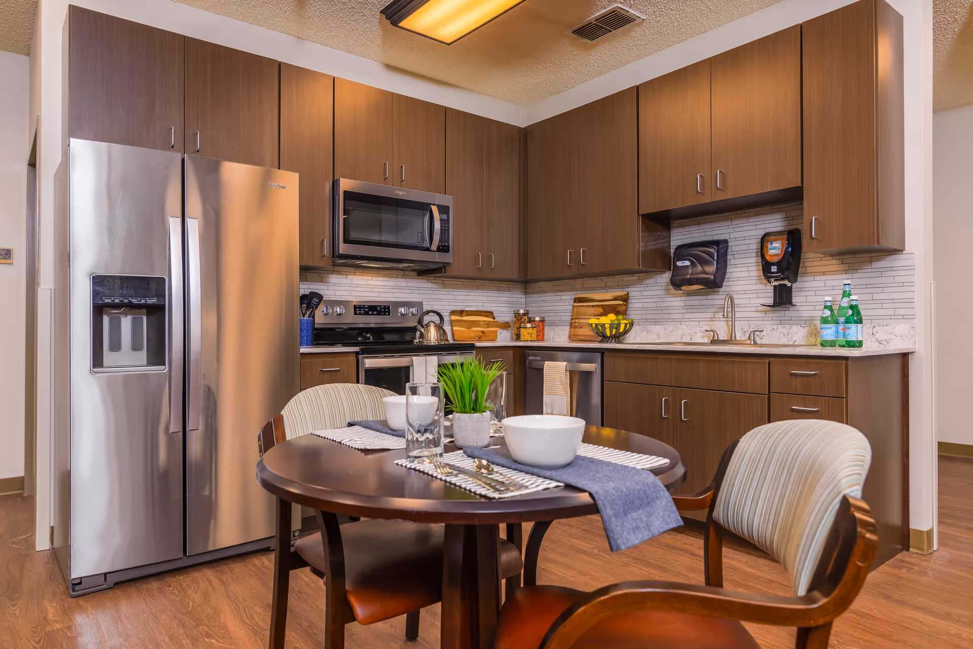 A modern kitchen and dining area with a round table set with bowls and glasses, wood cabinets, and stainless steel refrigerator and appliances.