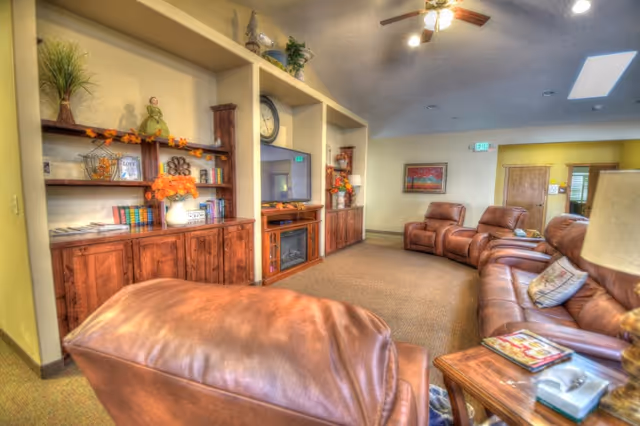 A cozy common living room with brown leather recliners arranged around a built-in entertainment unit with a TV, decorative shelving, and an electric fireplace.