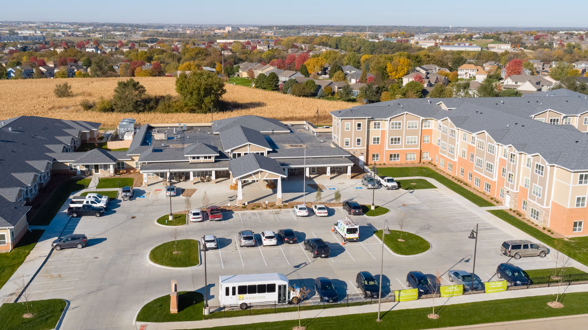 Aerial view of Cedarhurst Senior Living of La Vista facility showing a large parking lot with several cars and a shuttle bus, surrounded by multi-story residential buildings with gray roofs. The background includes a field and a suburban neighborhood with trees displaying fall colors under a clear sky.