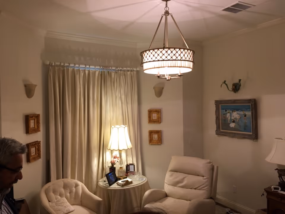 A cozy living room with cream-colored walls and curtains, featuring a round side table with a lamp and decorative items, two comfortable armchairs, framed pictures on the walls, and a ceiling light fixture. A man with gray hair and glasses is partially visible on the left side.