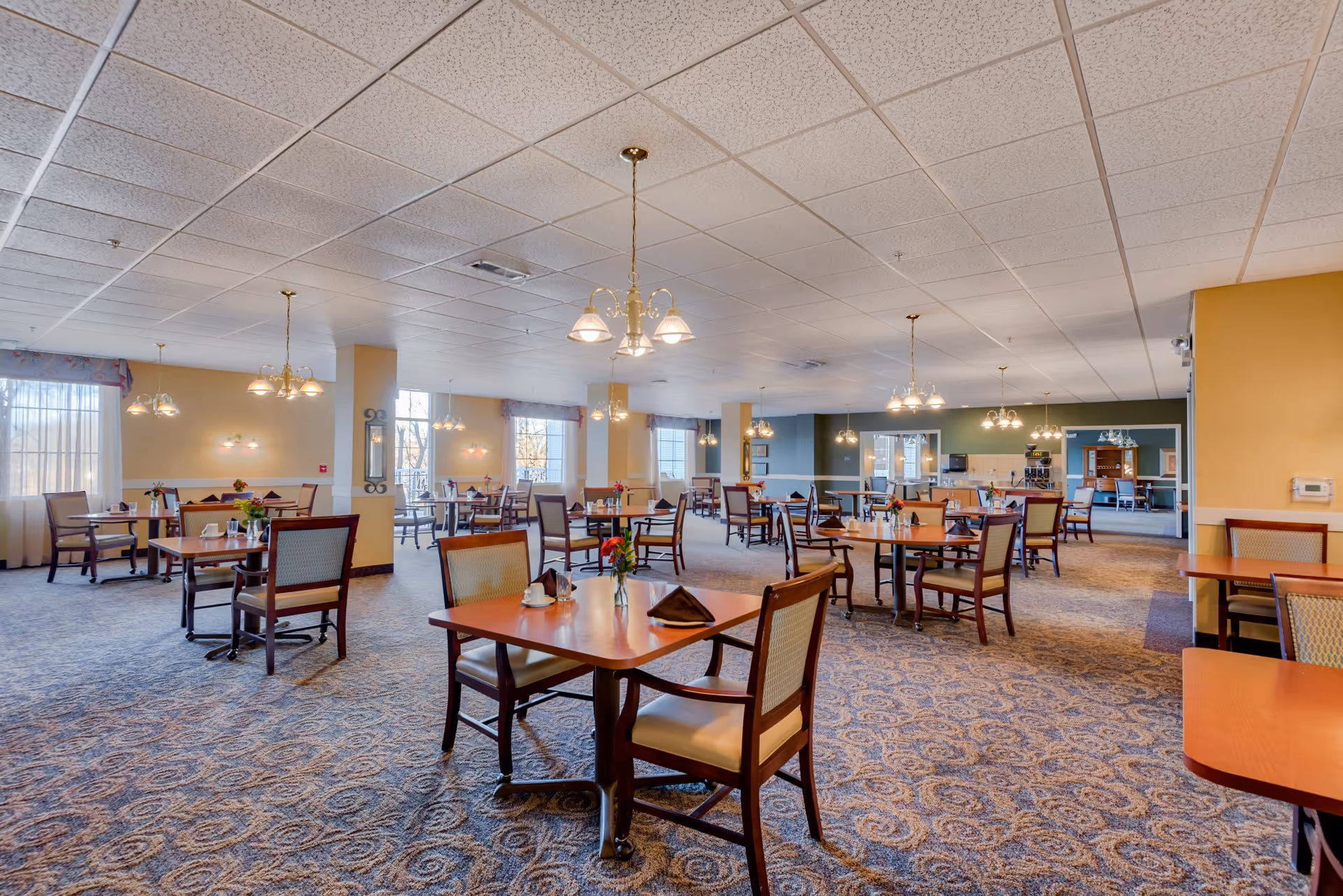 A spacious dining room with multiple wooden tables and chairs arranged neatly. The room has large windows with sheer curtains allowing natural light to fill the space. Elegant chandeliers hang from the ceiling, and the carpet has a patterned design. The walls are painted in soft yellow and green tones, creating a warm and inviting atmosphere.