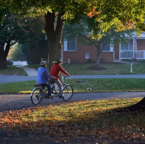 Two people wearing helmets ride a tandem bicycle down a tree-lined street in front of a brick house.