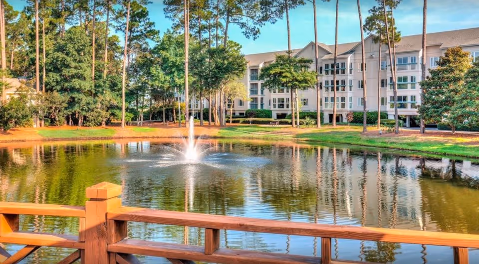 View across a pond with a central fountain, wooden railing in the foreground and a multi-story Tidepointe building surrounded by trees.