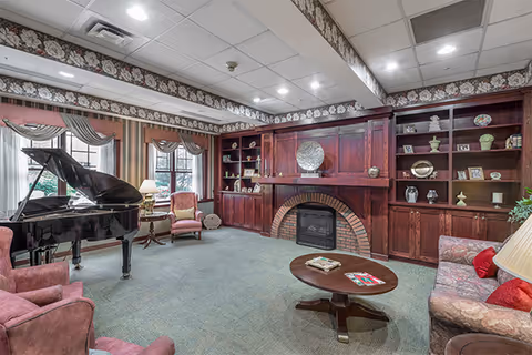 Cozy common room with a grand piano, upholstered chairs and sofa, a coffee table, and a wood-paneled fireplace with built-in shelves.