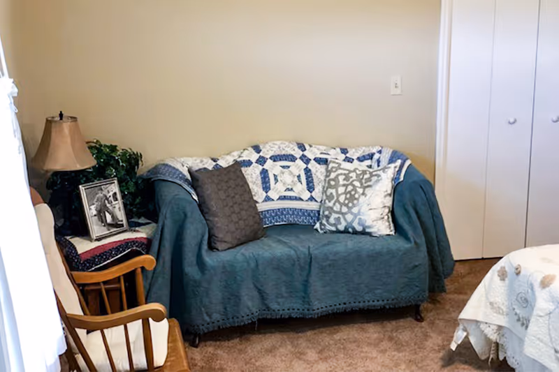 A cozy room with a small couch covered by a blue throw and two decorative pillows, a wooden rocking chair with a cushion, a side table with a lamp, a framed black and white photo, and a plant. There is a bed partially visible on the right side with a white quilt, and a closed white closet door in the background.