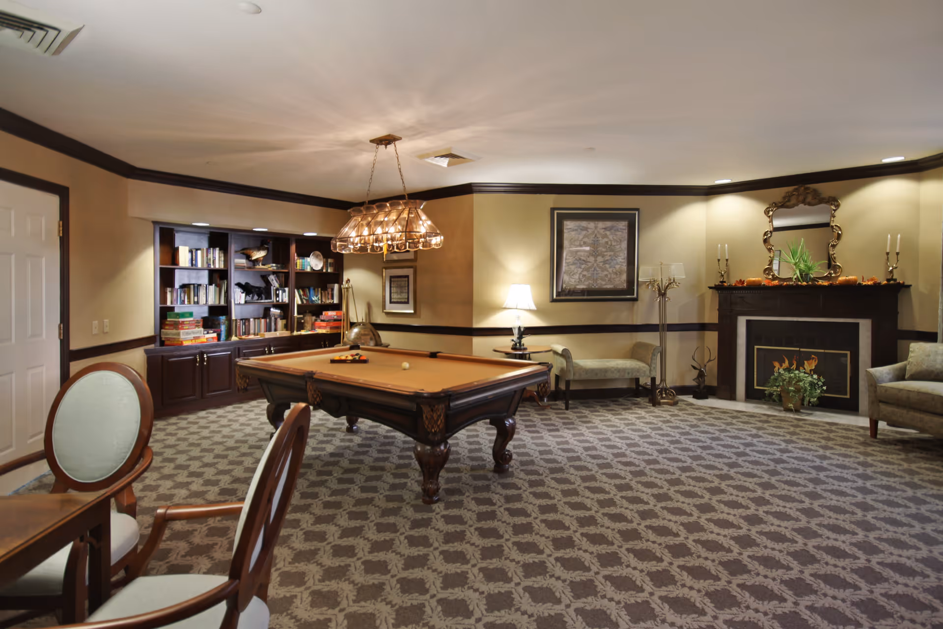 A cozy game room with a pool table in the center, surrounded by upholstered chairs and a bench. The room features a patterned carpet, a bookshelf filled with books and board games, a lit fireplace with decorative items on the mantel, and warm lighting from a hanging chandelier and table lamps.