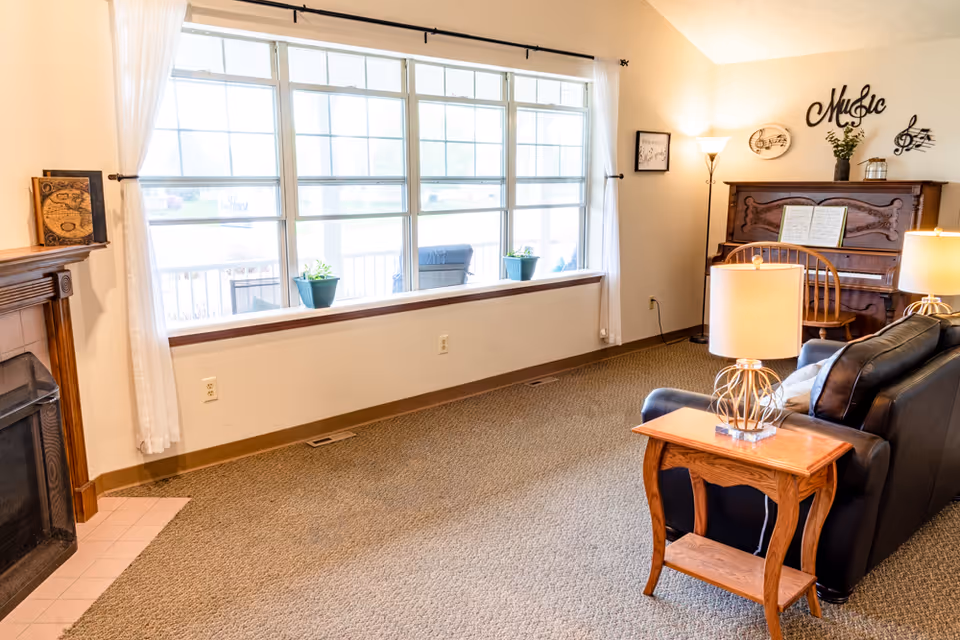 A cozy living room with a large window covered by white curtains, a black leather sofa, a wooden side table with a lamp, a wooden piano with sheet music and musical decorations on the wall, and a fireplace on the left side.