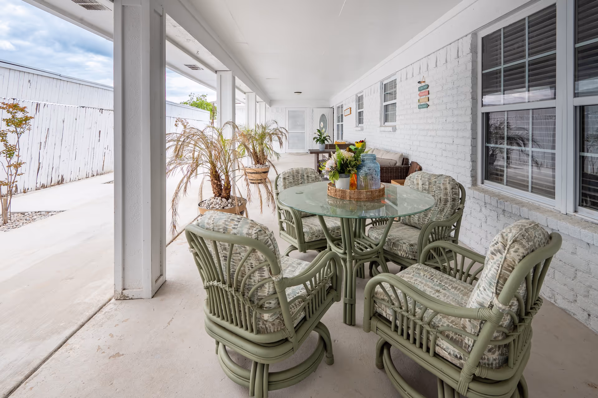 Covered outdoor patio area with a round glass table surrounded by five cushioned green wicker chairs. There are potted plants along the white brick wall and a white door at the far end. The patio overlooks a concrete pathway with a white wooden fence on the left side.