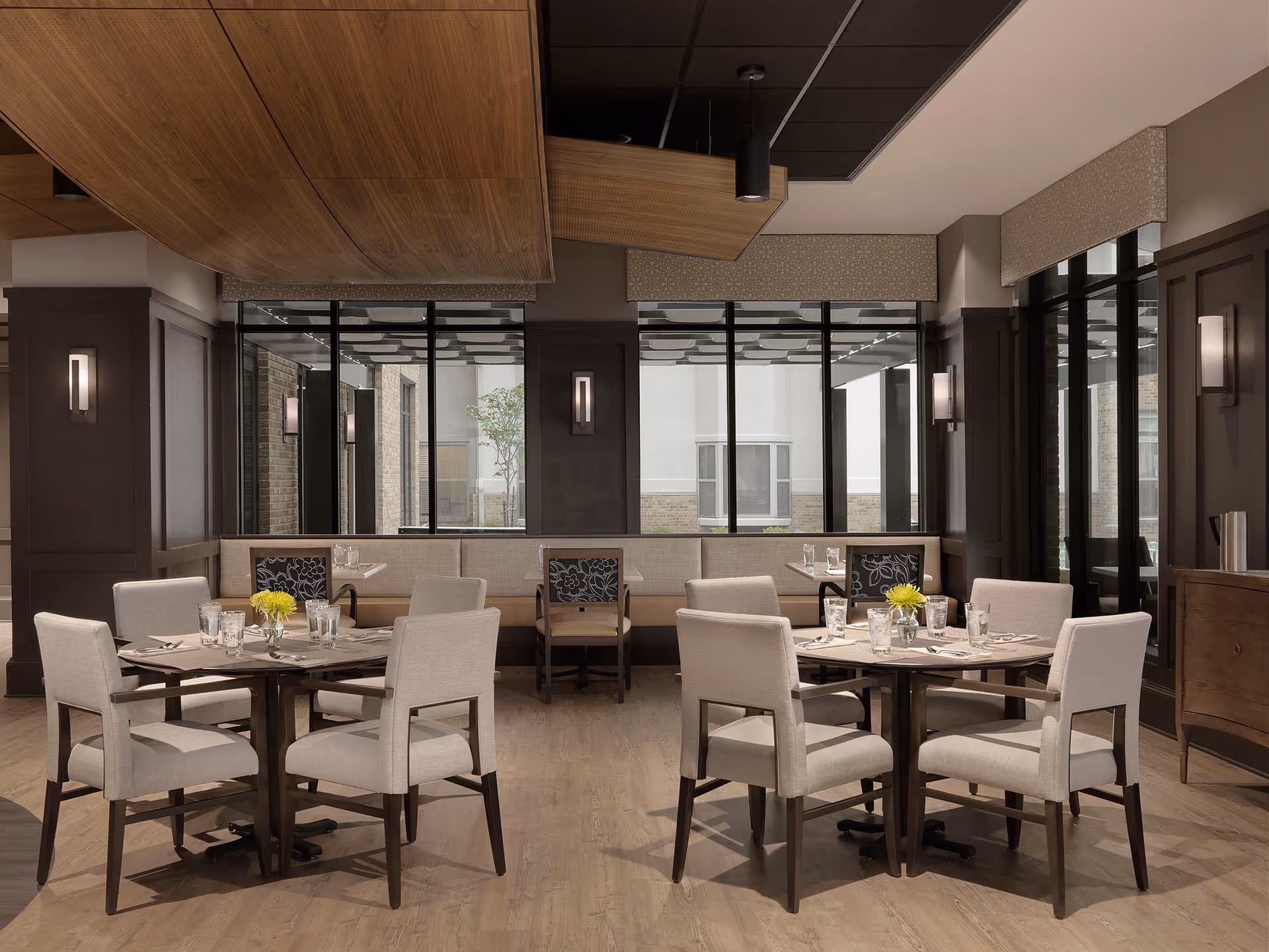A modern dining room in a senior living facility with two round tables, each surrounded by four beige upholstered chairs. The tables are set with glasses, silverware, and small vases with yellow flowers. Large windows with black frames line the back wall, allowing natural light to enter. The room features wood flooring, dark wood paneling on the walls, and a unique wooden ceiling design.