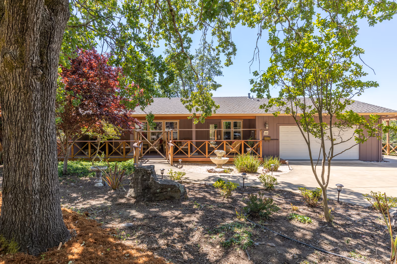 Front exterior of a single-story ranch-style building with a wooden porch, garage, fountain, and trees in a landscaped yard.