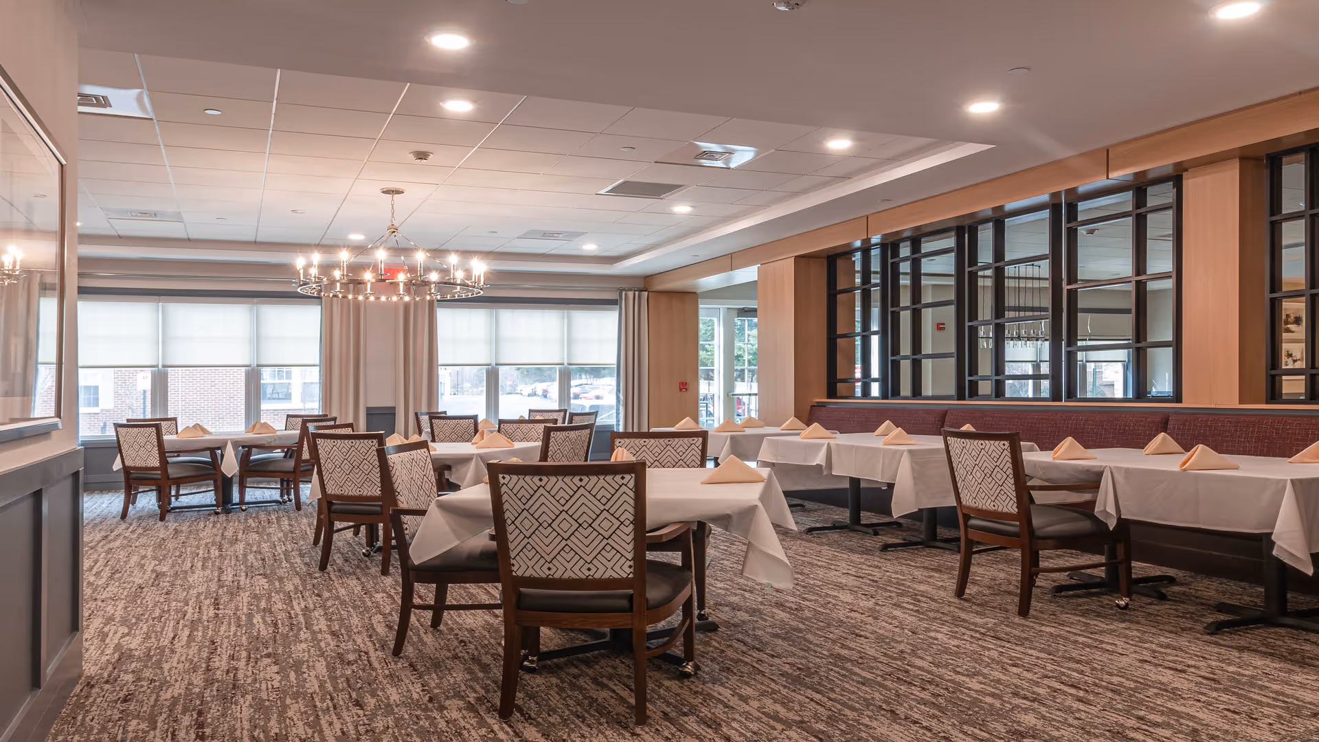 A spacious dining room with multiple tables covered in white tablecloths and neatly folded napkins. The room features patterned chairs, large windows with blinds, a chandelier, and a carpeted floor. There is a mirrored wall with wooden framing on one side.