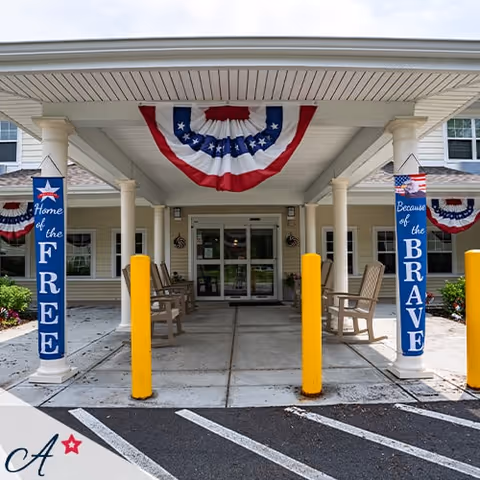 Entrance of a senior living facility decorated with patriotic red, white, and blue bunting. Two pillars display banners reading 'Home of the FREE' and 'Because of the BRAVE'. There are chairs on either side of the entrance under a covered walkway with yellow bollards in front.