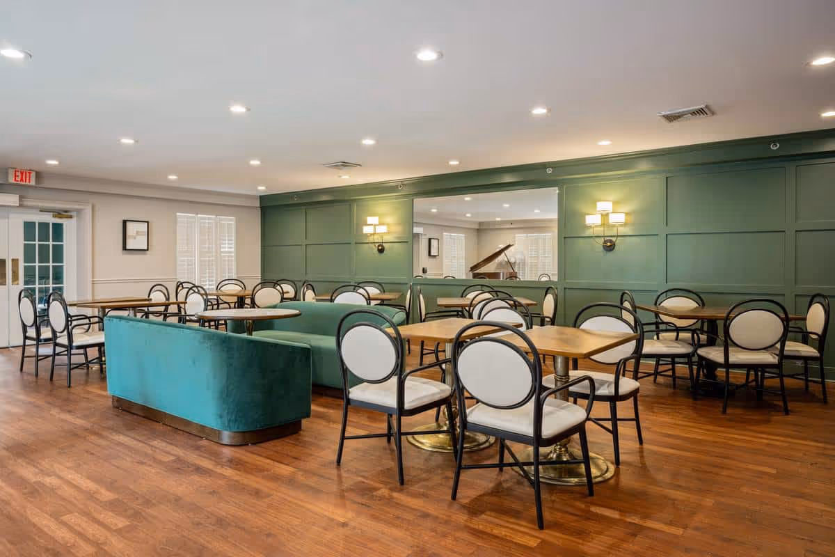 A spacious dining room with wooden floors, multiple tables and chairs with white cushioned seats and backs, and a green upholstered bench. The walls are painted green with decorative paneling and wall sconces providing warm lighting. There is a large mirror on one wall reflecting a grand piano in an adjacent room.