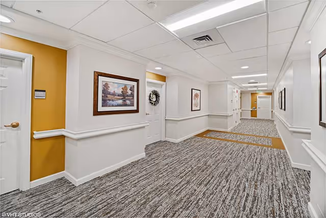 Well-lit carpeted interior hallway with white walls, handrails, framed artwork and several doors.