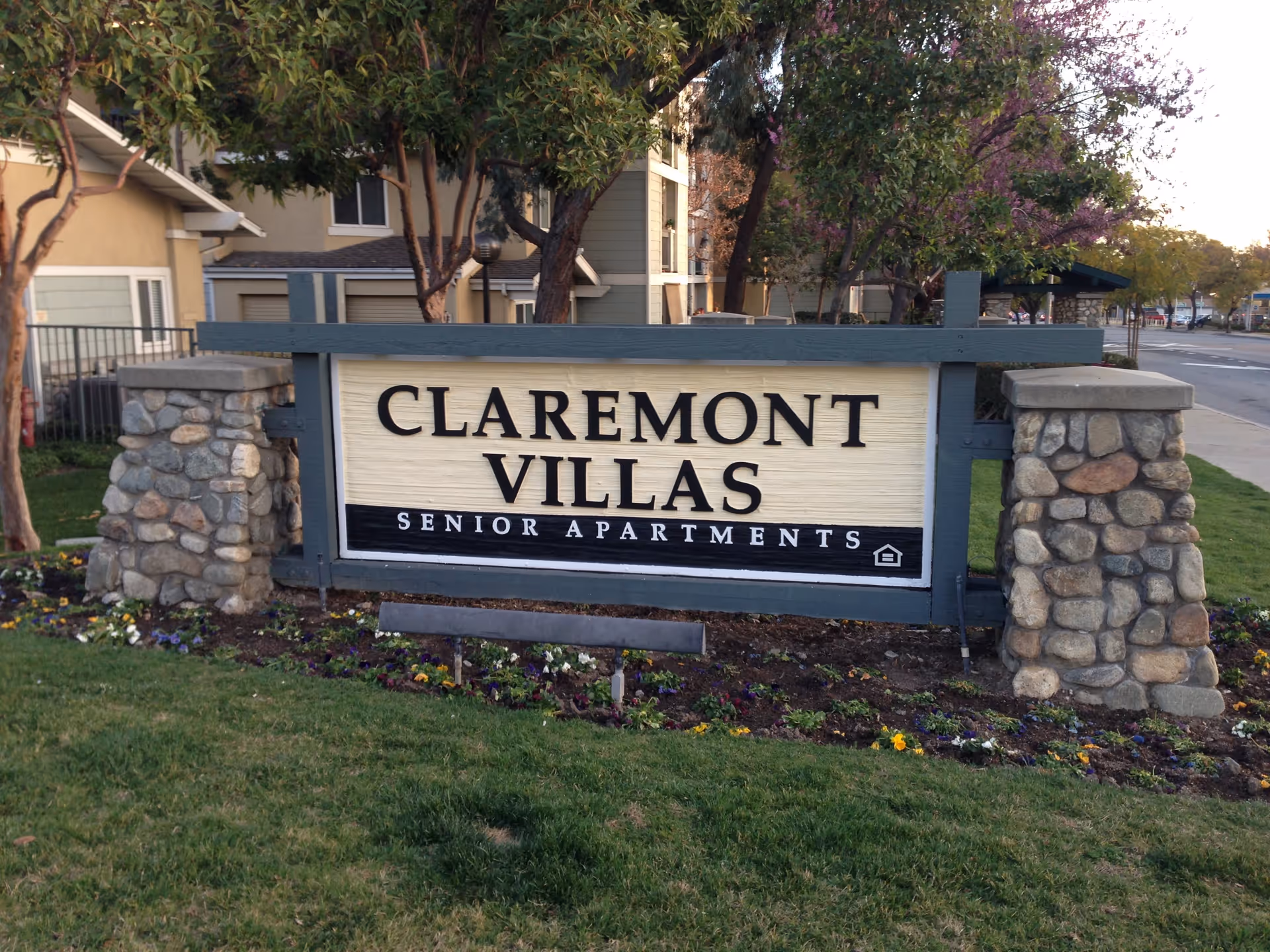 Entrance sign reading "Claremont Villas Senior Apartments" flanked by stone pillars with apartment buildings and trees in the background.
