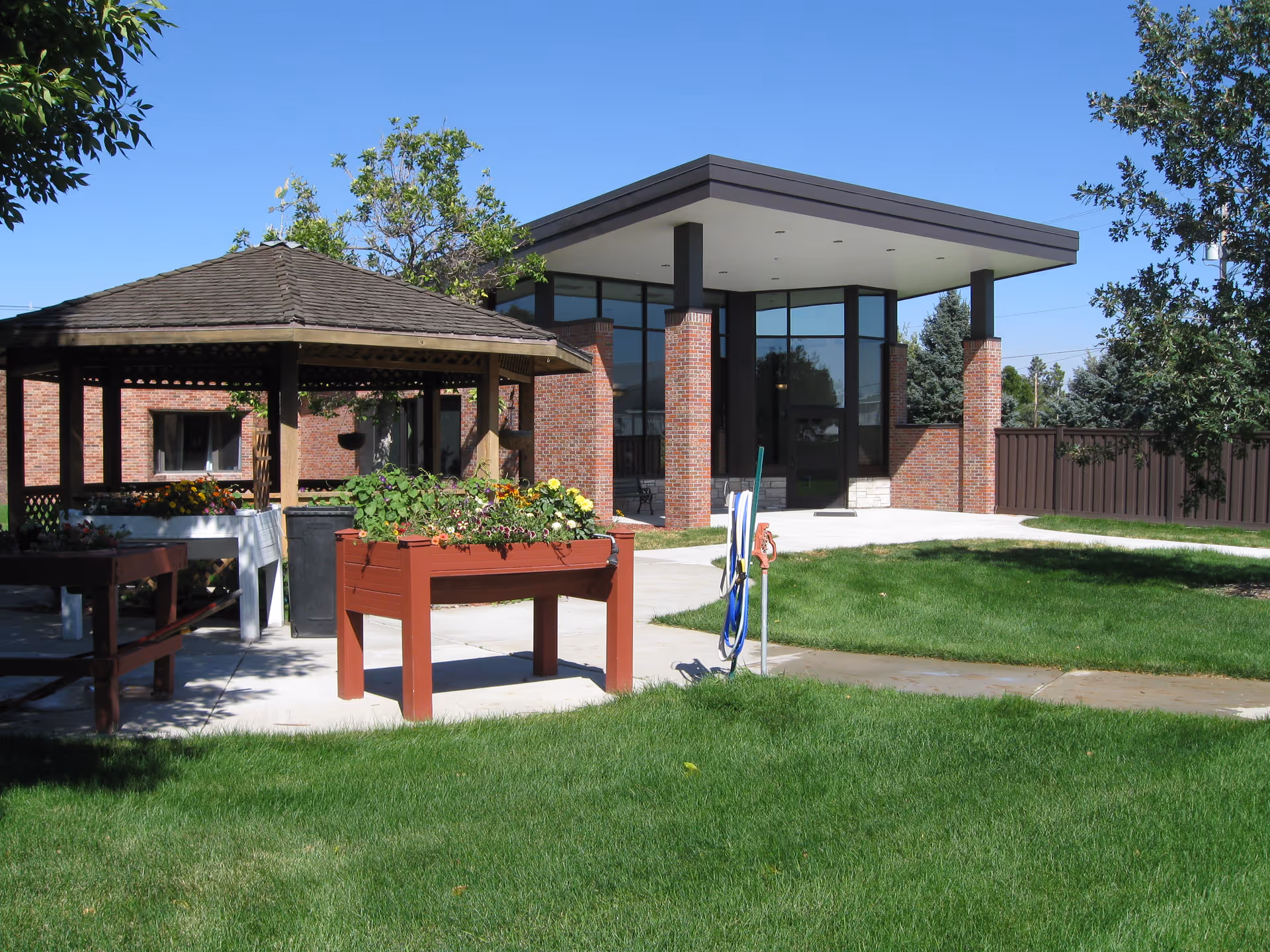 A landscaped courtyard with a wooden gazebo, raised planter boxes, and the brick entrance of the facility under a clear blue sky.