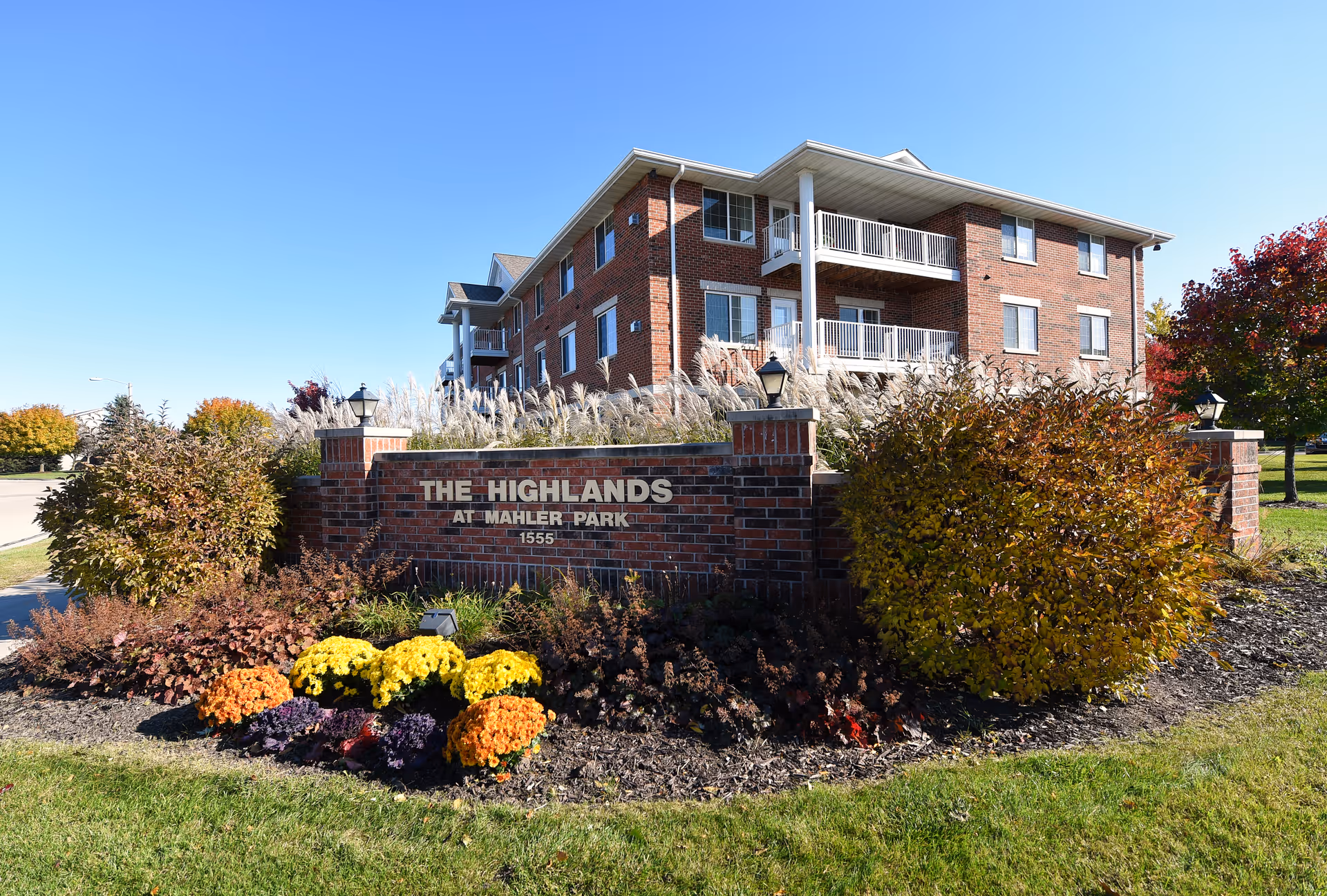 Exterior view of a three-story brick apartment building with balconies at The Highlands at Mahler Park Apartments 55+. In front of the building is a brick sign with the facility name and address, surrounded by landscaped bushes and colorful flowers under a clear blue sky.