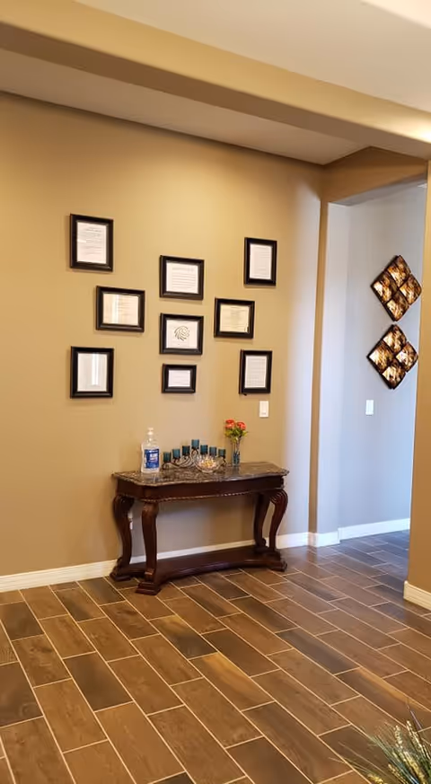 A hallway corner in an assisted living facility with a wooden console table against a beige wall. The table holds a bottle of hand sanitizer, a decorative menorah, a small bowl, and a small vase with red flowers. Above the table, there are nine framed certificates or documents arranged in a grid pattern. The floor is covered with brown wood-like tiles, and there is a wall opening leading to another room with decorative wall art visible.