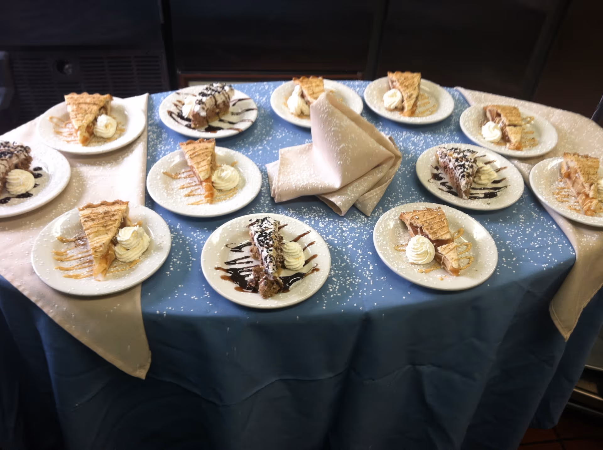 A round table covered with a blue tablecloth and a beige runner, displaying multiple plates of dessert slices with whipped cream and drizzled sauces, dusted with powdered sugar. A folded beige napkin is placed in the center of the table.
