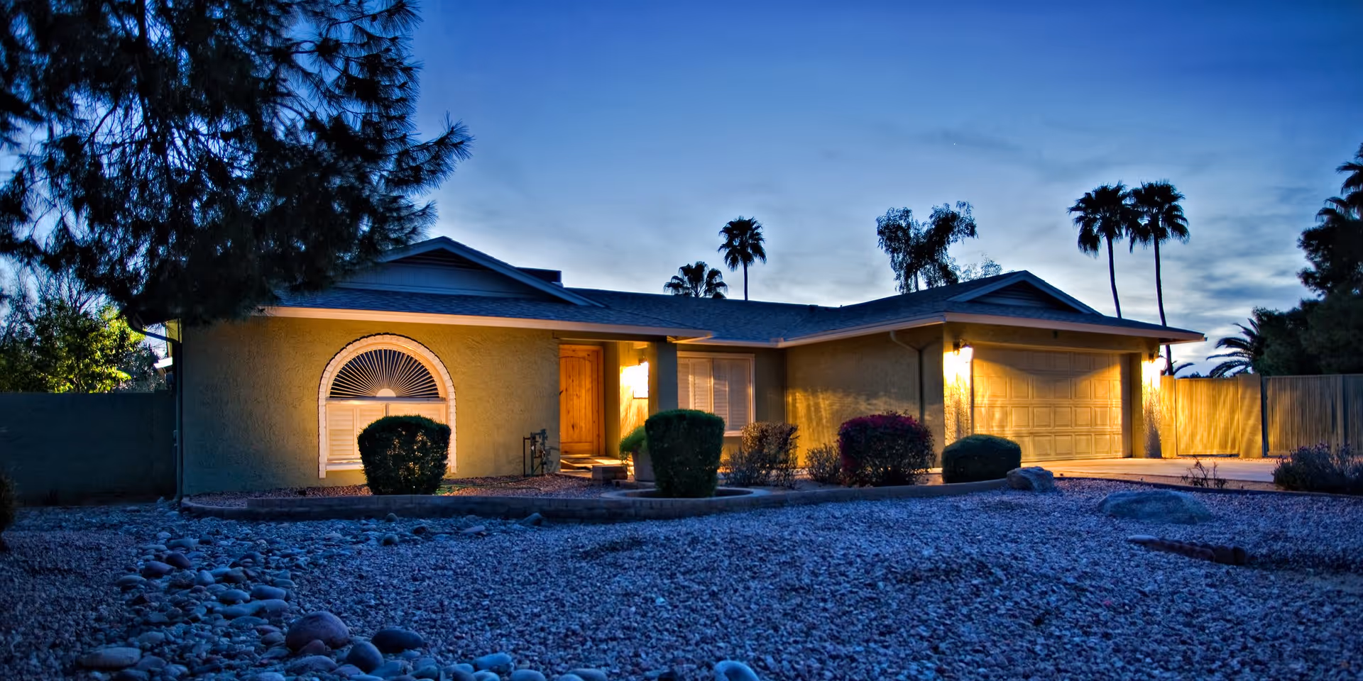 Exterior view of a single-story house at dusk with lights illuminating the front entrance and garage. The yard is covered with gravel and has several bushes and trees, including palm trees in the background.