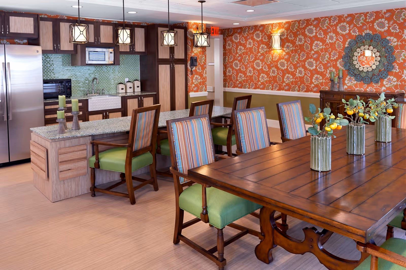Bright communal dining area with a long wooden table, striped and green chairs, and an open kitchen with an island and pendant lights.