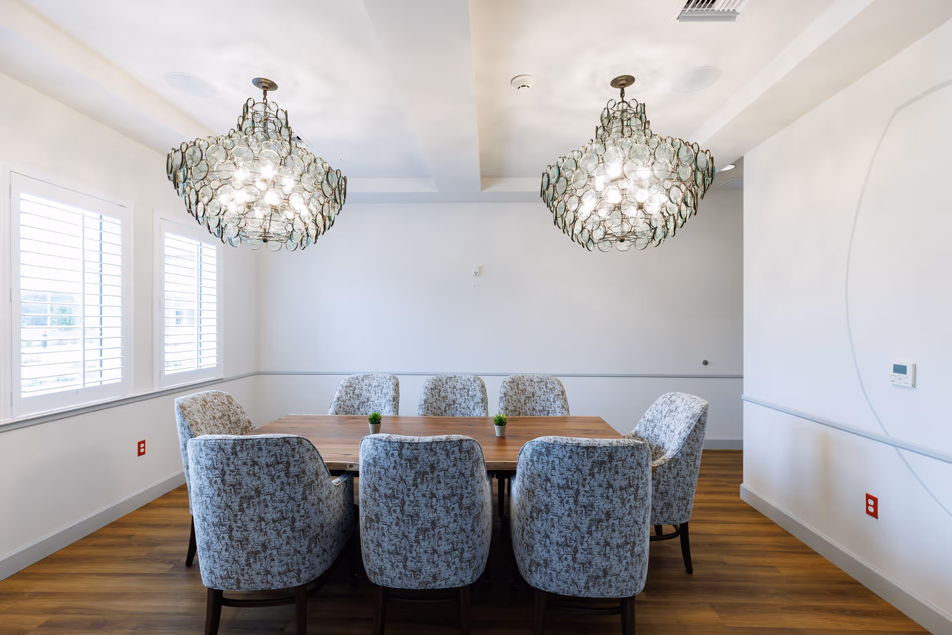 A bright dining room with a wooden table surrounded by eight upholstered chairs with a floral pattern. Two large, decorative chandeliers hang from the ceiling. The room has white walls, three windows with white shutters, and a wooden floor.