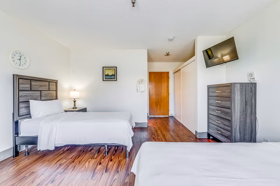 A bright and clean bedroom in an assisted living facility with two single beds made with white linens. The room features wood flooring, a wooden dresser with a flat-screen TV mounted above it, a nightstand with a lamp, a wall clock, and a framed picture on the wall. There is a wooden door and a closet with sliding doors.