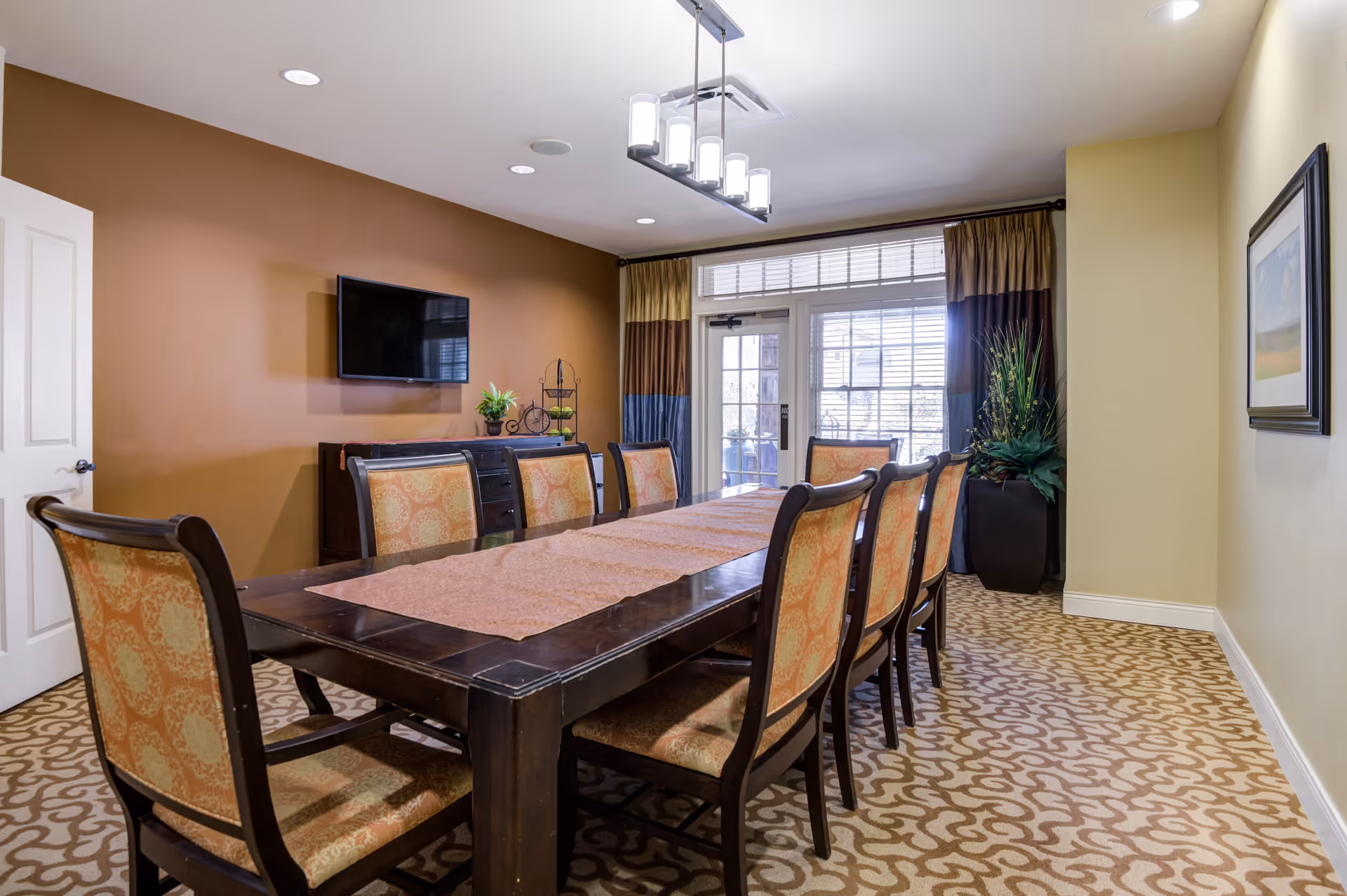 A dining room with a long dark wooden table covered by a brown table runner and surrounded by eight upholstered chairs with patterned fabric. The room has a patterned carpet, a brown accent wall with a mounted flat-screen TV, a sideboard with decorative items, and a large window with glass doors covered by curtains. A modern ceiling light fixture hangs above the table.