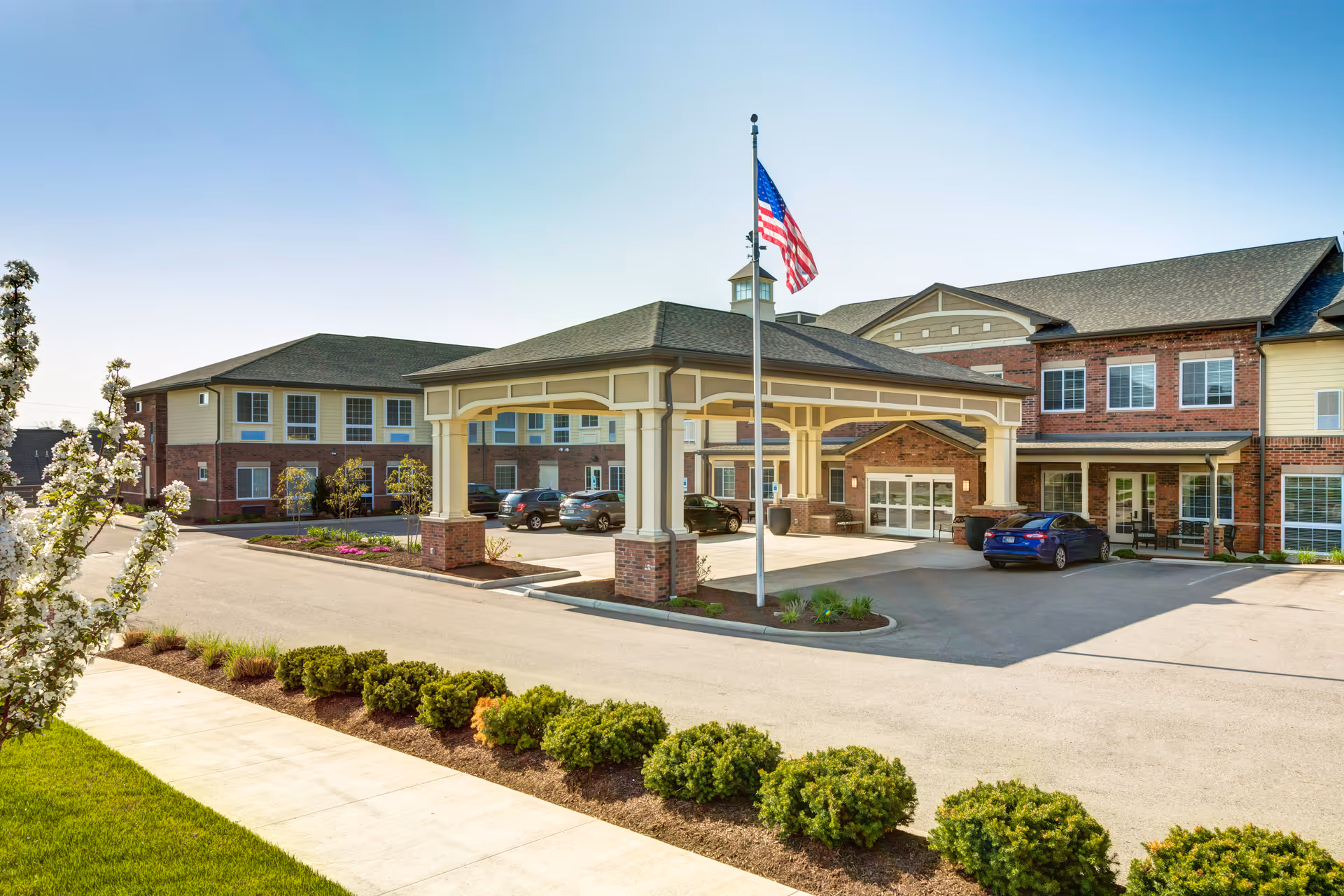 Exterior view of Traditions of West Chester senior living facility showing a large covered entrance with an American flagpole in front, surrounded by a parking area with several cars and landscaped greenery.