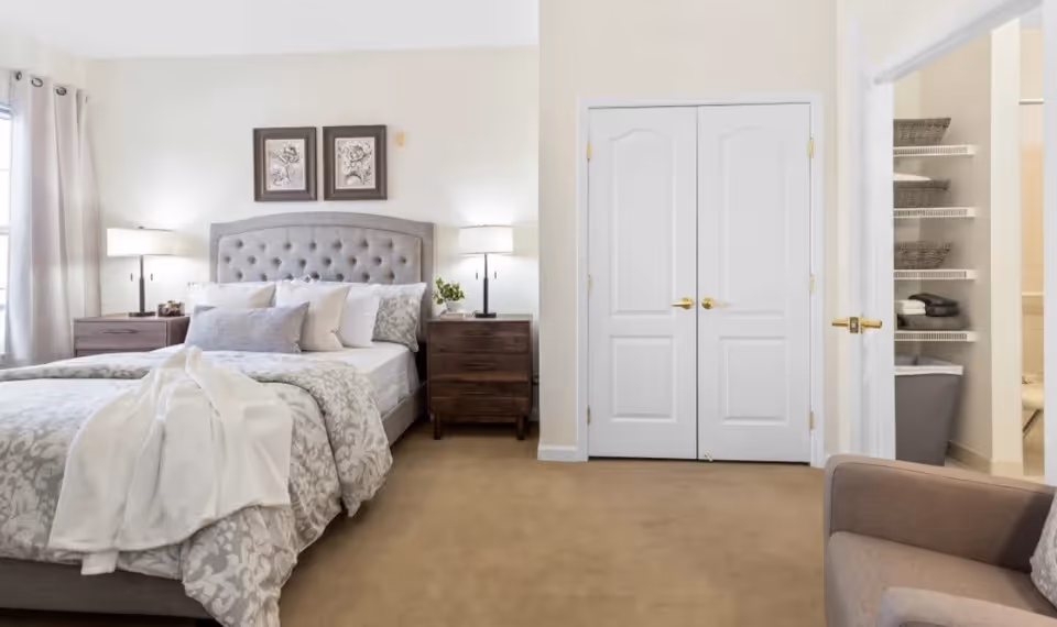 A neatly made bed with a tufted gray headboard and multiple pillows in a bedroom. There are two dark wooden nightstands with lamps on either side of the bed. To the right, there is a double-door closet and an open doorway showing shelves with baskets and folded towels. A beige armchair is partially visible in the foreground on the right side.