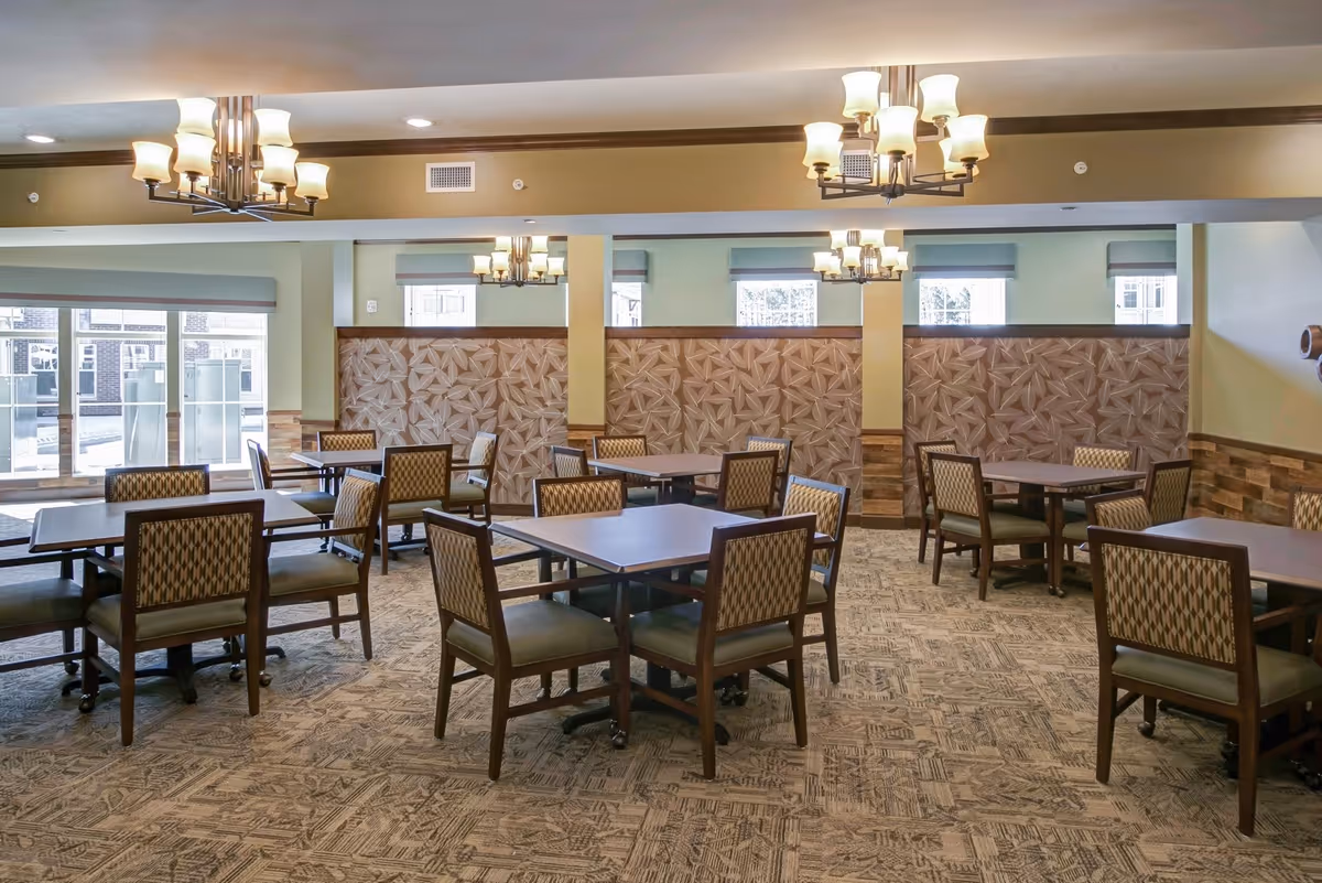 A spacious dining room with multiple square tables and cushioned chairs arranged neatly. The room features patterned carpet flooring, decorative wall panels, large windows letting in natural light, and ceiling chandeliers providing warm lighting.