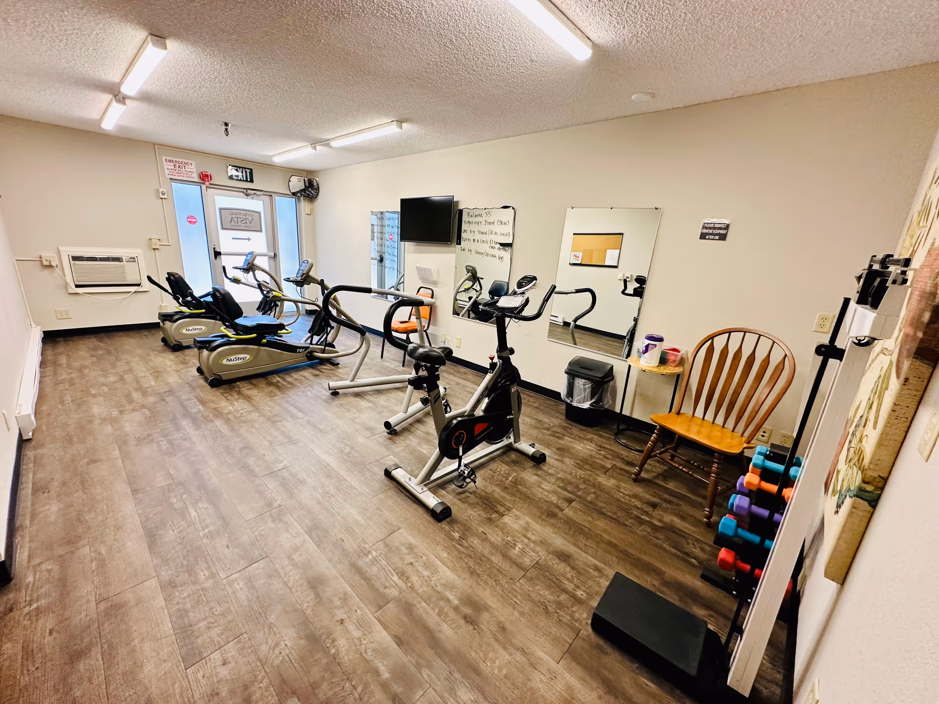A small fitness room with exercise equipment including two recumbent bikes, an upright stationary bike, and a treadmill. The room has wood-look flooring, white walls, a wall-mounted TV, a mirror, a wooden chair, and a rack of colorful dumbbells. There is an emergency exit door with a glass window and an air conditioning unit on the wall.
