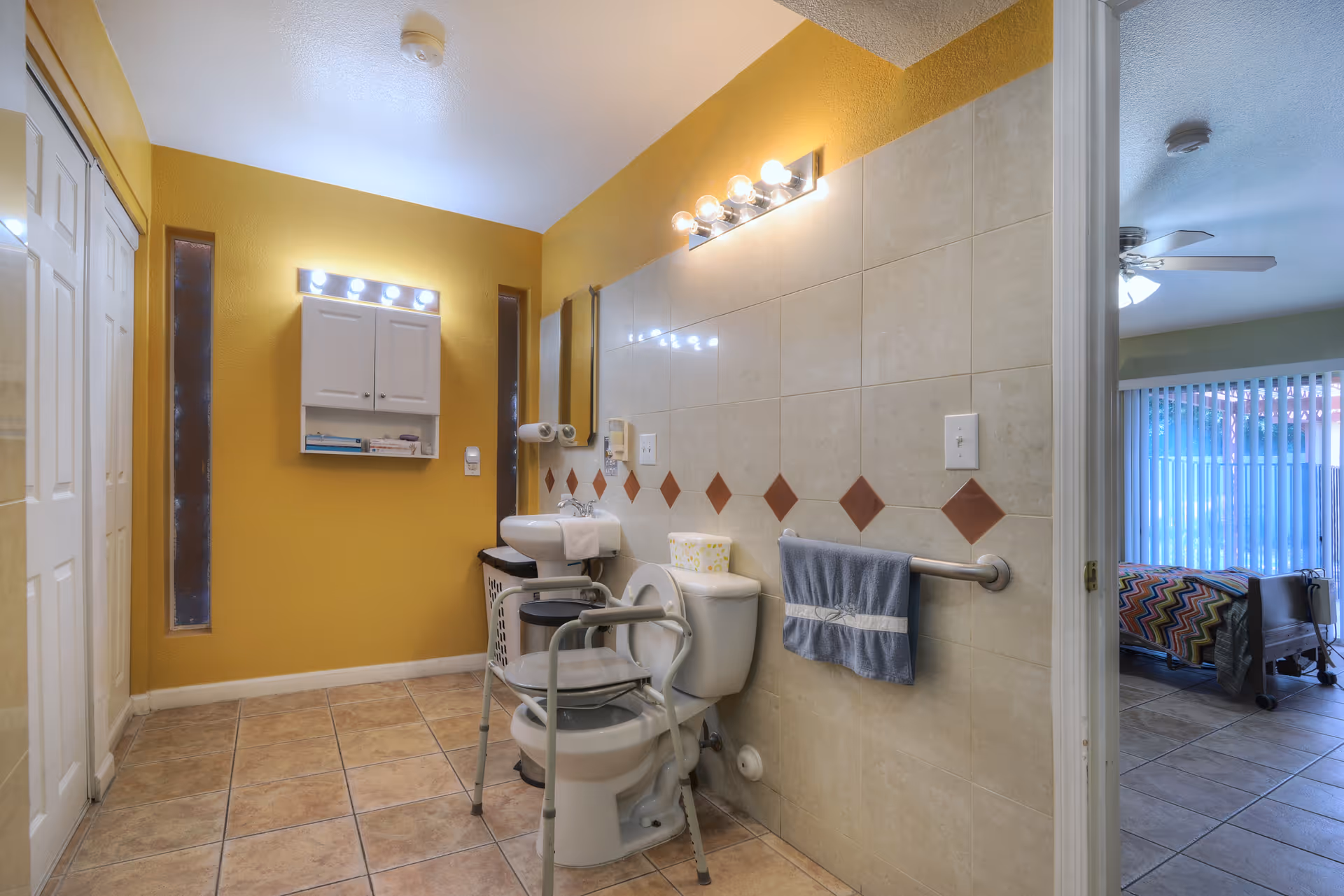 A bathroom with yellow walls and tiled floor featuring a toilet equipped with a safety frame, a small sink with a mirror and light above it, a towel rack with a blue towel, and a wall-mounted cabinet. The bathroom opens into a room with a bed and a ceiling fan, visible through the doorway.