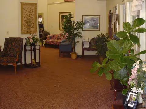 Indoor senior living lounge with chairs, plants, and framed artwork on carpet.