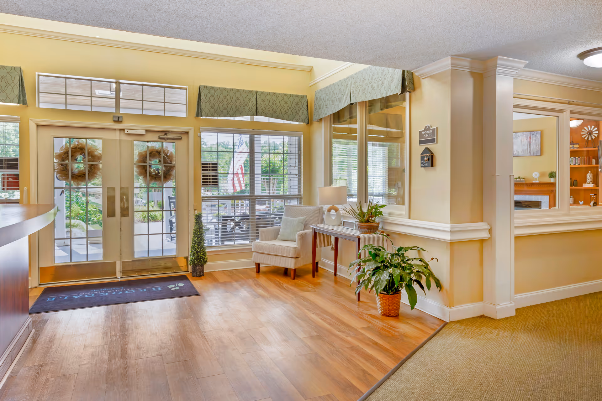 Welcoming senior living facility lobby with double glass doors, seating area, potted plants, and wood flooring.