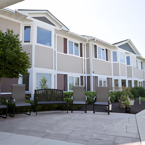 Outdoor patio area with several chairs and benches arranged along the side of a two-story building with multiple windows. There are bushes and small trees planted near the building, and the sky is clear.