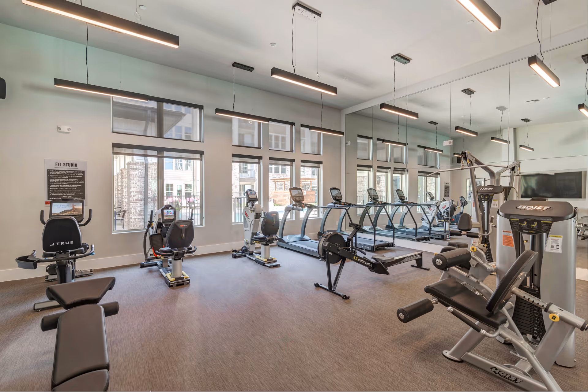 A bright and modern fitness studio with various exercise equipment including stationary bikes, treadmills, a rowing machine, and weight machines. Large windows allow natural light to fill the room, and a wall of mirrors reflects the equipment and space. The room has hanging linear lights and a carpeted floor.