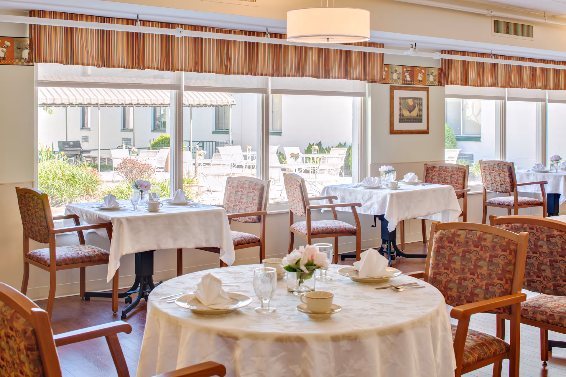 A bright dining room with round and square tables covered with white tablecloths, set with plates, cups, glasses, and napkins. Upholstered chairs with patterned fabric surround the tables. Large windows provide a view of an outdoor patio area with tables, chairs, and greenery.