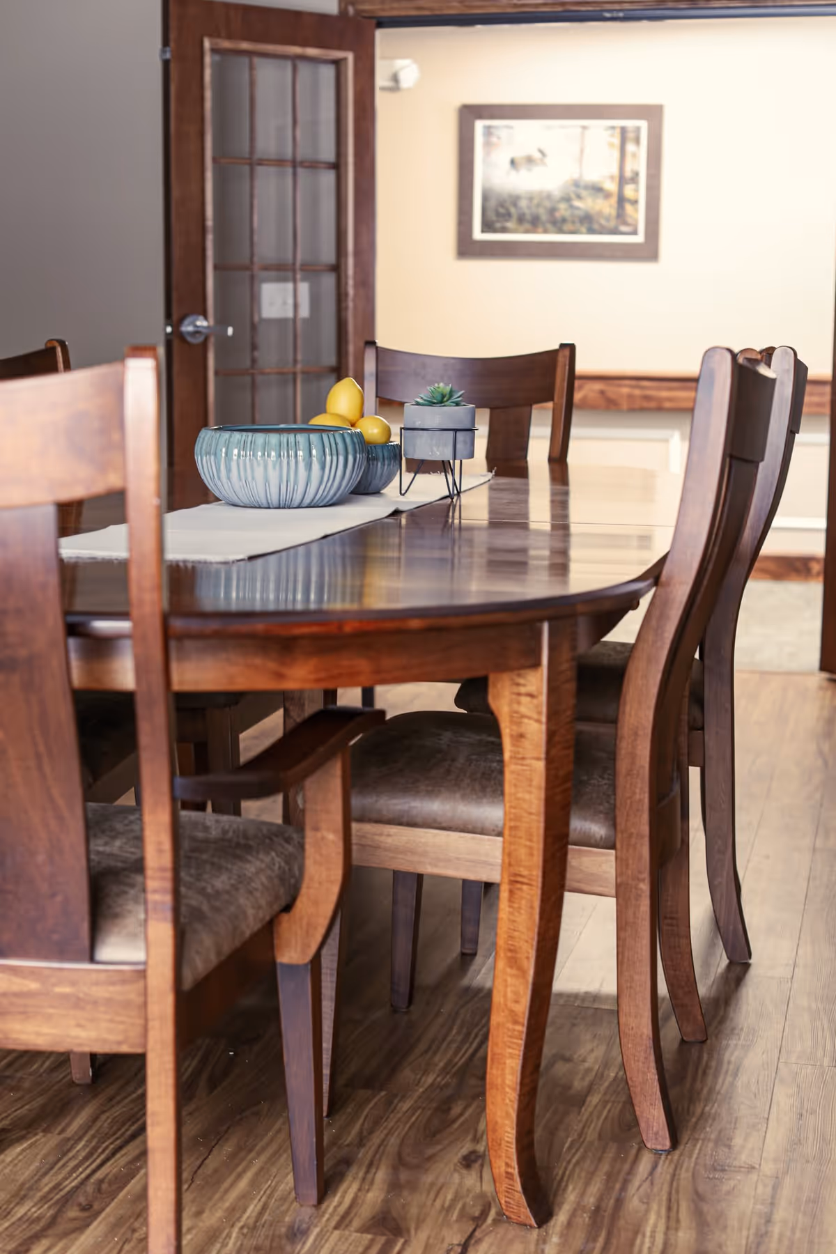 A wooden dining table with matching wooden chairs arranged around it. On the table, there is a decorative blue bowl with lemons and a small potted succulent plant. In the background, there is a wooden door with glass panels and a framed picture on a beige wall.