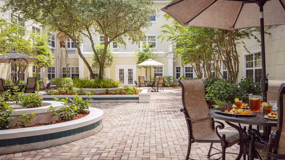 Outdoor courtyard area with brick paving, round tables with umbrellas, chairs, and greenery including trees and shrubs. A table in the foreground has pitchers and glasses of iced tea along with plates of fruit and snacks. The courtyard is surrounded by a multi-story building with many windows.