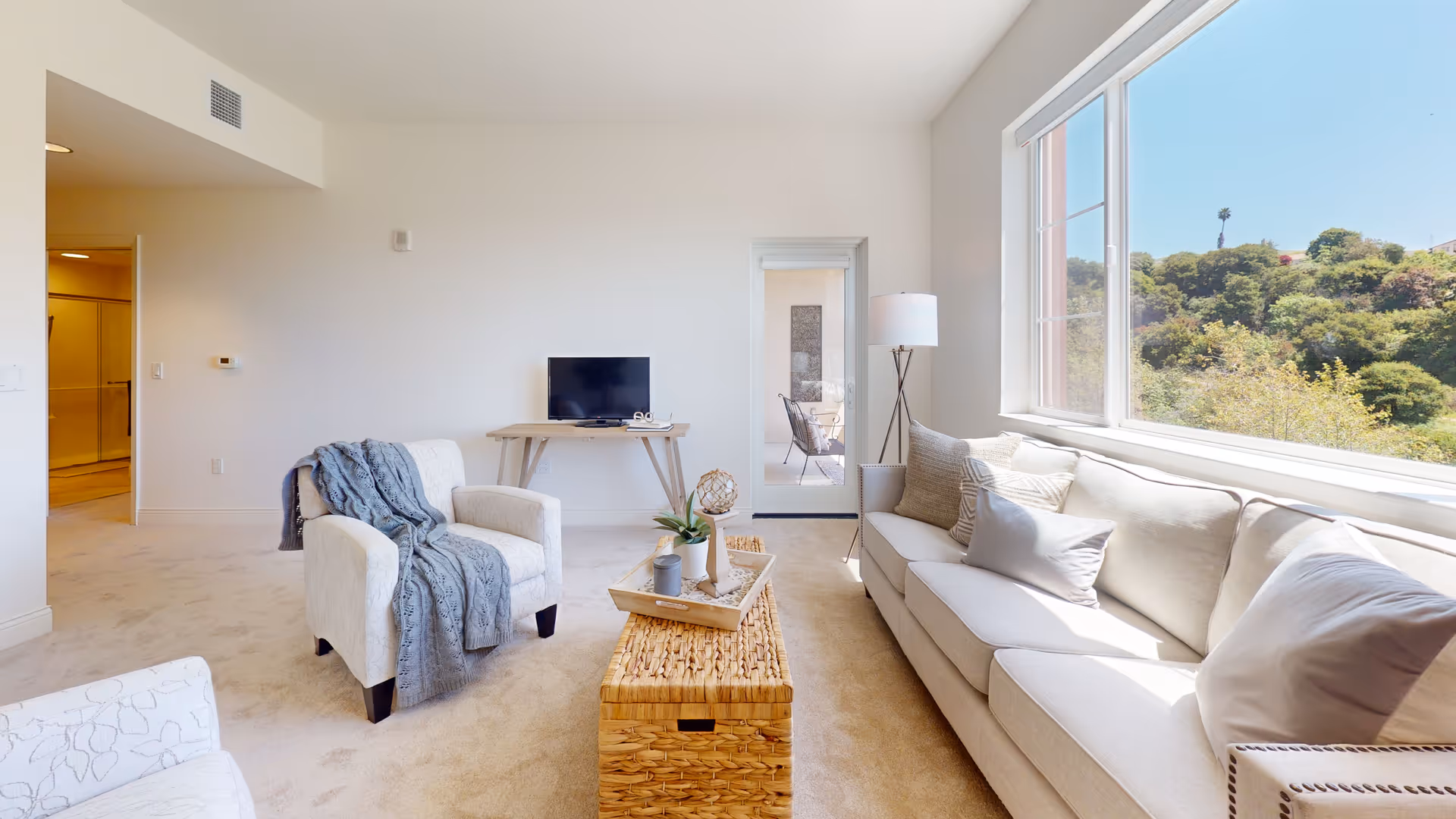 A bright and spacious living room with a large window showing a view of green hills outside. The room features a beige sofa with multiple cushions, a white armchair draped with a gray knitted throw, a woven coffee table with decorative items, a small wooden table with a flat-screen TV, and a tall floor lamp. The walls are white, and the carpet is light beige. A glass door leads to another room with additional seating.