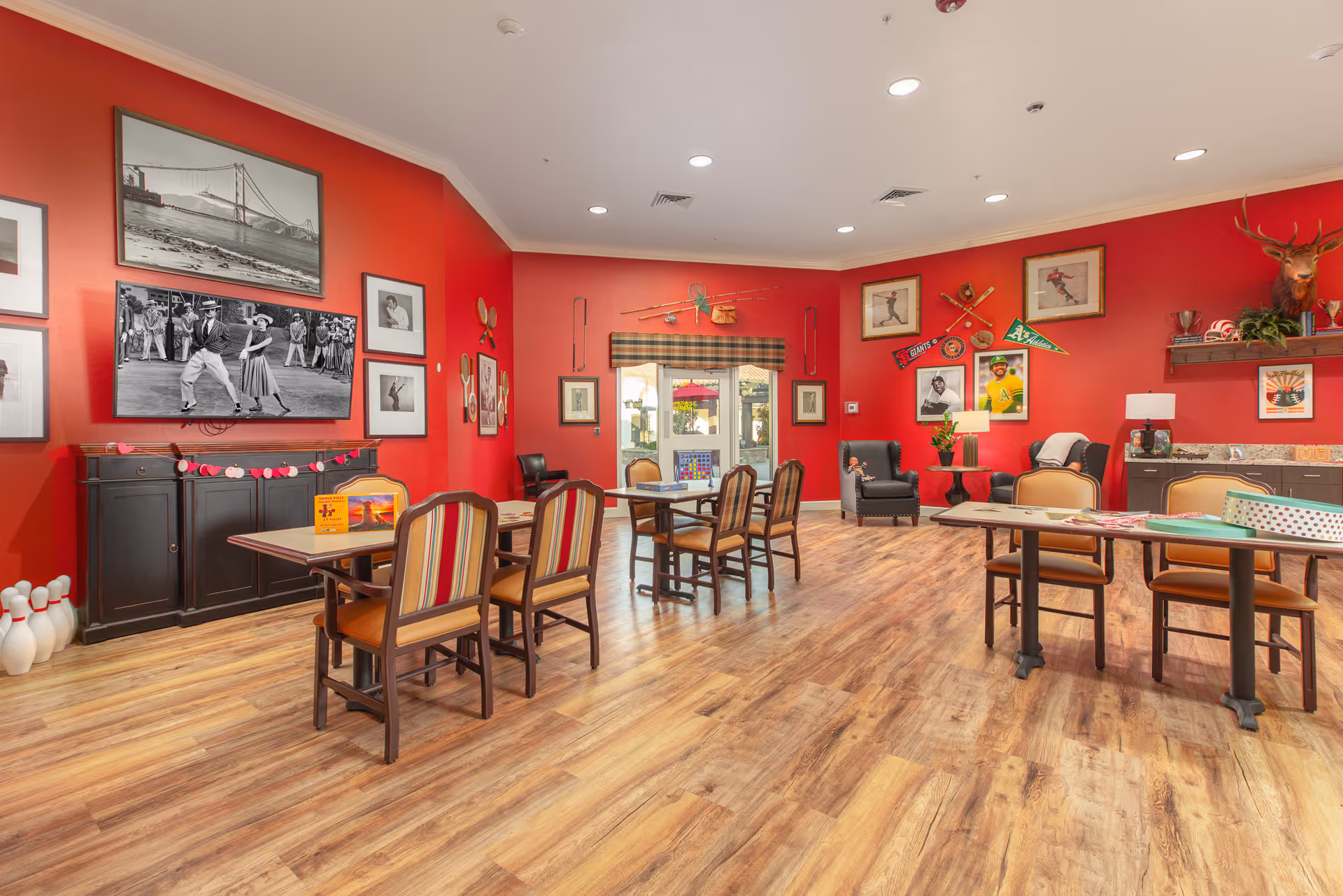 A brightly lit common room with red walls and wood flooring featuring several tables and chairs arranged for group activities. The walls are decorated with framed pictures, sports memorabilia, and a mounted deer head. There is a cabinet with a large black and white photo above it and a set of bowling pins on the floor. Large windows with a plaid valance let in natural light.