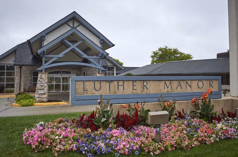 Exterior view of the entrance to Luther Manor, featuring a stone and wood building with a peaked roof and a large sign in front surrounded by colorful flowers.
