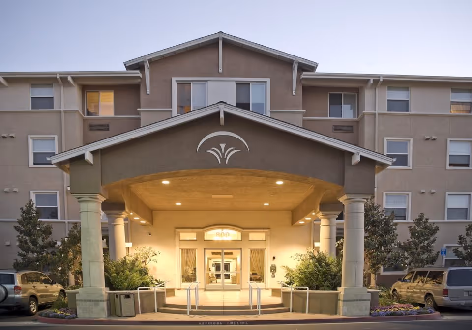 Front entrance of a multi-story senior living facility named Heritage Estate Senior Apartments, featuring a covered driveway with columns, well-lit entryway, and surrounding landscaping with bushes and trees. Several parked cars are visible on either side of the entrance.