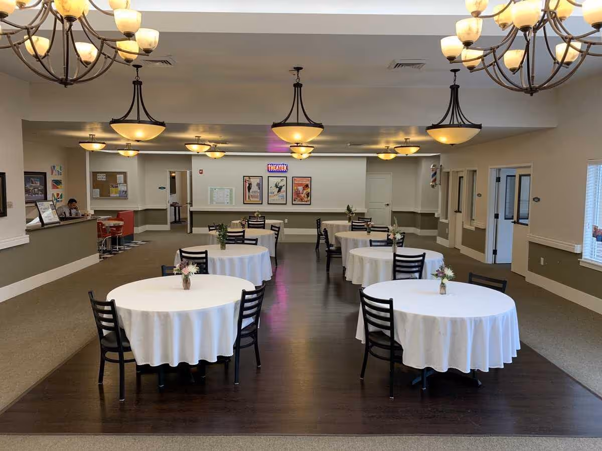 A dining area with round tables covered in white tablecloths, each table decorated with a small vase of flowers. The room has dark wood flooring in the center and carpet around the edges. Multiple hanging light fixtures illuminate the space. There is a reception desk on the left side with a person sitting behind it, and framed posters on the far wall with a neon sign that reads 'THEATER'. Several doors and windows are visible along the walls.