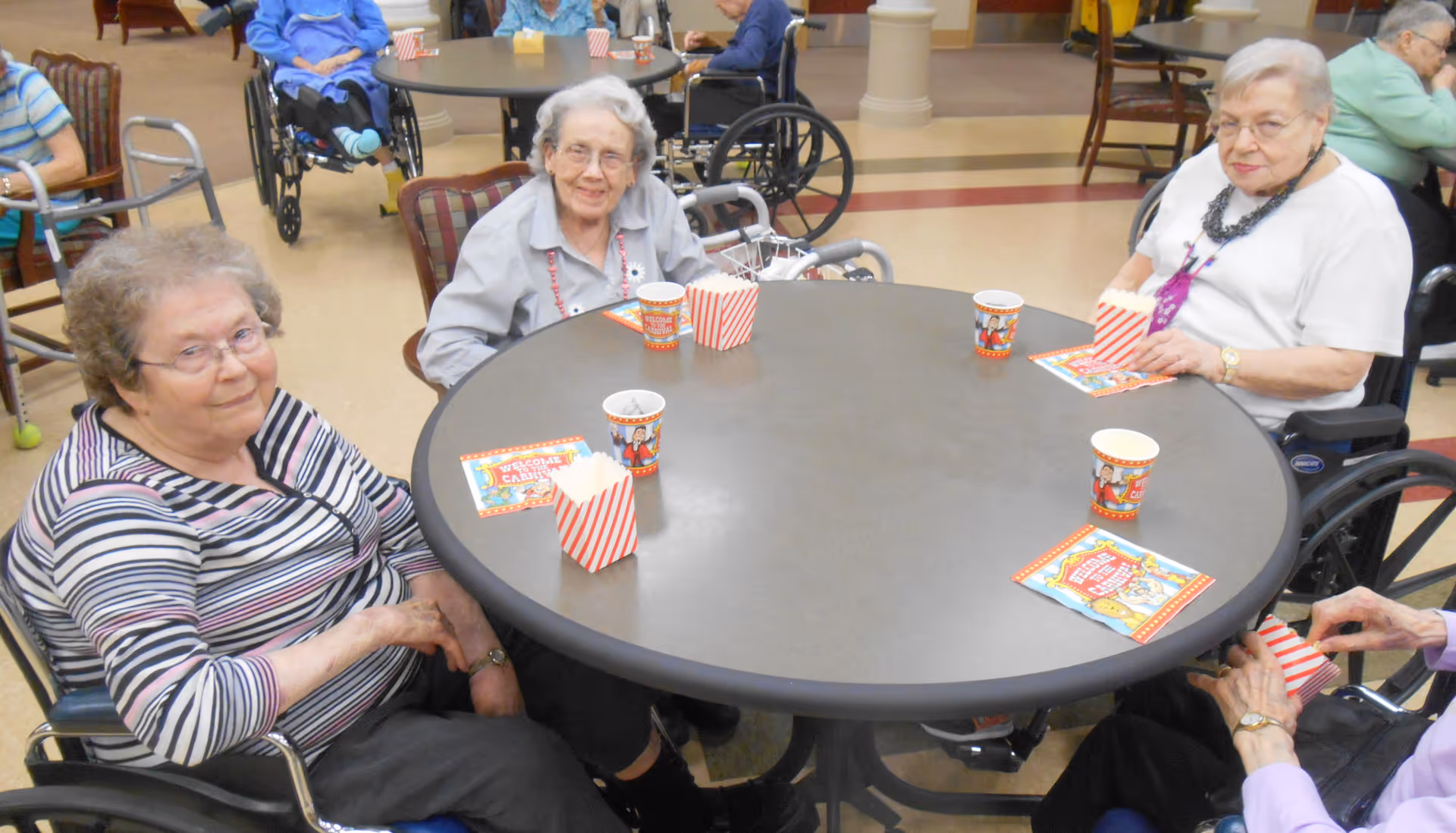 A group of elderly women sitting around a round table in a communal area, each with a small popcorn container and a themed paper cup, likely participating in a social or recreational activity.