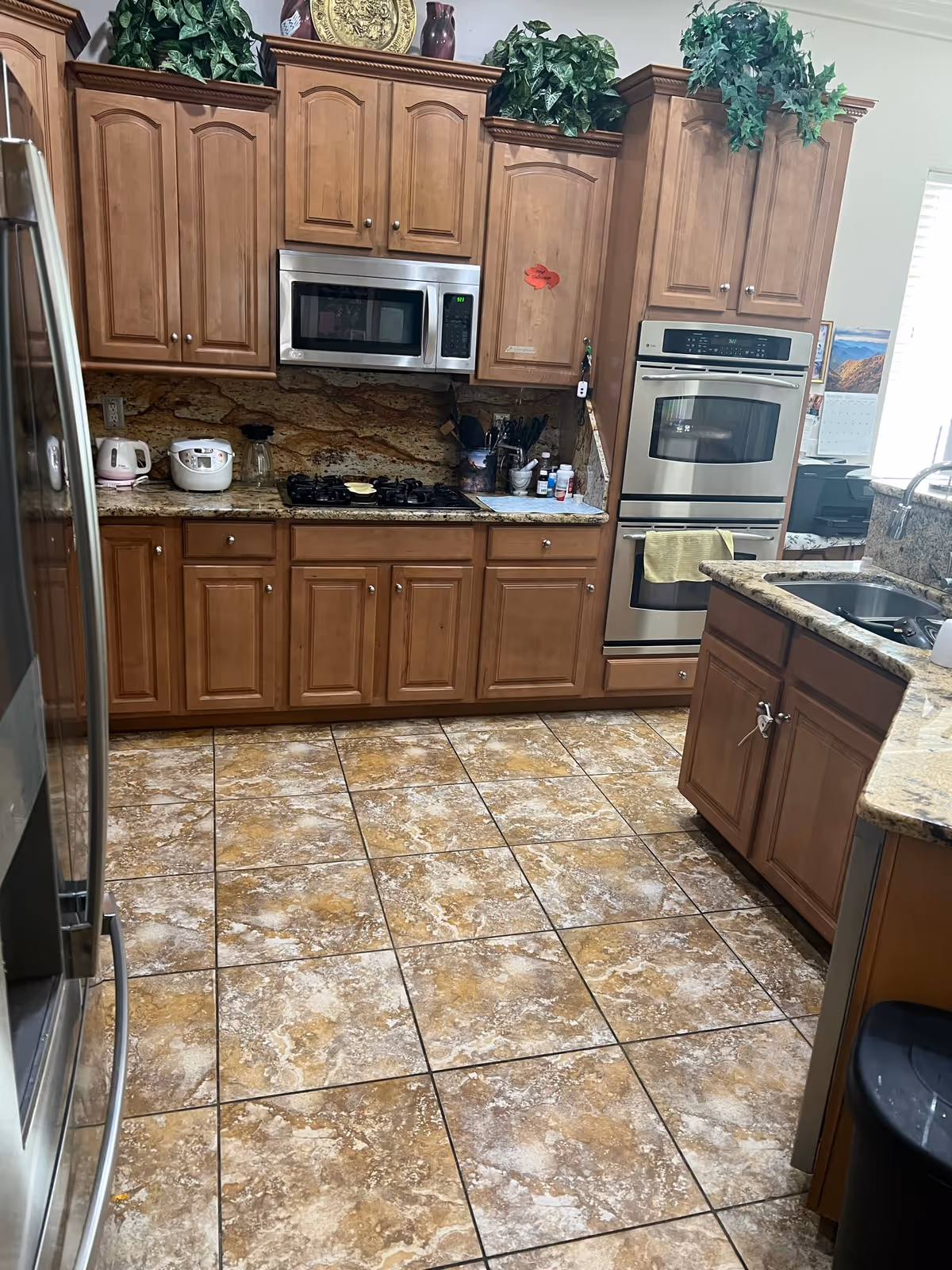 A kitchen with wooden cabinets, a stainless steel microwave above a gas stove, a double wall oven, and a granite countertop with a sink. The floor is tiled with brown and beige tiles. There are various kitchen appliances and utensils on the countertop, and decorative plants on top of the cabinets.