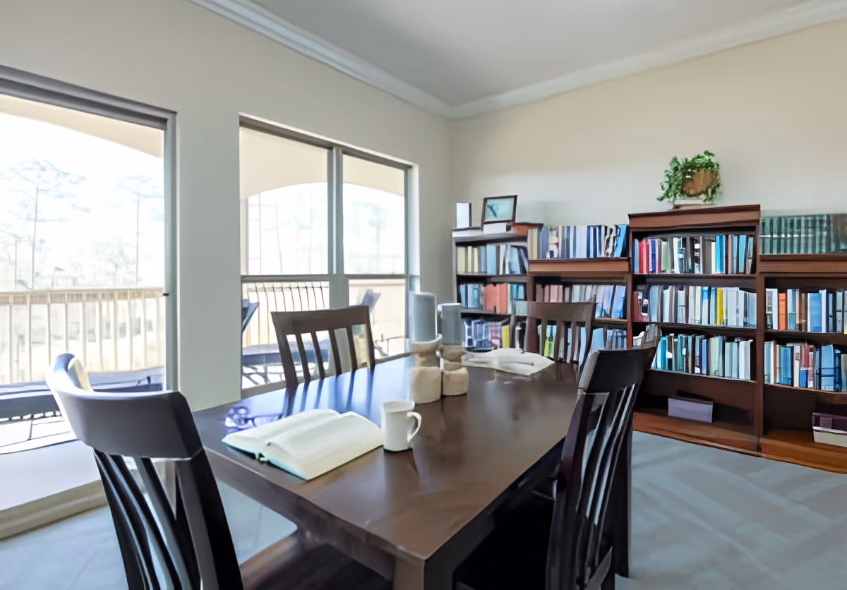 Bright communal dining and reading room with a wooden table and chairs, bookshelves along the wall, and large windows opening onto a balcony.